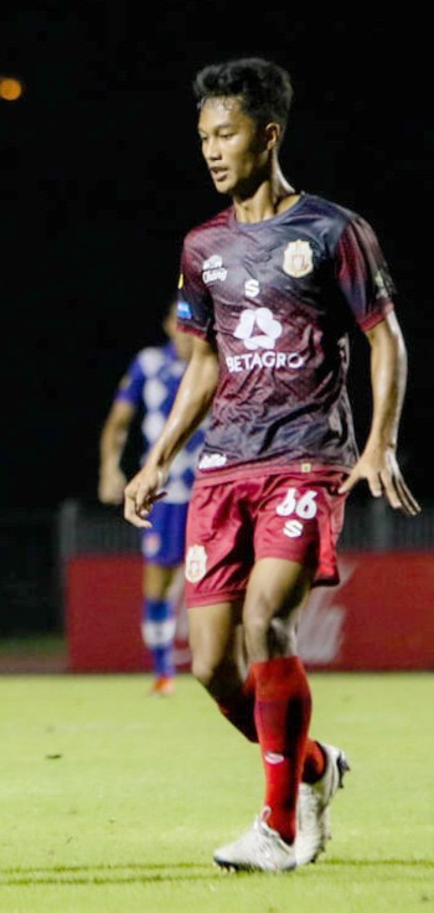 Ein Mann in einem maronenfarbenen Hemd und roten Shorts, der auf einem Feld Fußball spielt, mit einer anderen Person im Hintergrund und einer Tafel mit Text.