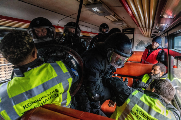 Polizisten in Einsatzausrüstung sitzen auf einem Bus, mit einer Person in der Mitte und einem Plakat im Hintergrund.