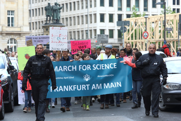 Eine Gruppe von Menschen marschiert mit einem "March for Science Frankfurt am Main"-Schild die Straße entlang, mit Autos, Gebäuden, Statuen, Laternenmasten, Schildern und Bäumen im Hintergrund.