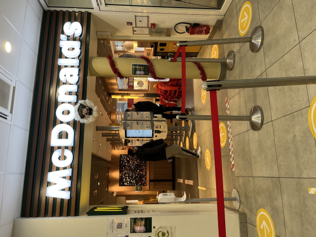 Innenseite eines McDonald's-Restaurants mit einem sichtbaren Schild, Metallstangen mit roten Bändern und einigen Menschen vor dem Eingang.