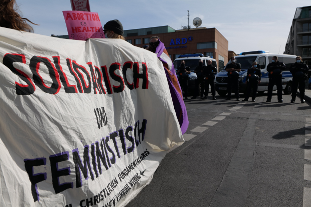 Eine Gruppe von Menschen marschiert auf einer Straße, hält ein Banner mit der Aufschrift 'Solidarität und Feminismus', mit parkenden Fahrzeugen und Gebäuden im Hintergrund unter einem klaren blauen Himmel.