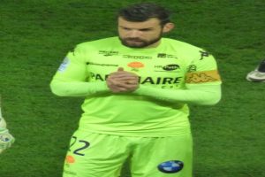 Ein Mann in einem gelben Torwarttrikot, der eine grüne T-Shirt und Shorts trägt, steht auf einem Fußballfeld und klatscht in die Hände, wobei auf der linken Seite des Bildes ein weiteres Bein zu sehen ist.