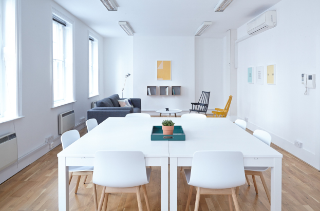 Innenraum eines Apartments in Berlin mit einem weißen Tisch mit einer Topfpflanze in einem Tablett, Stühlen, einer Couch mit Kissen, einer Lampe, Regalen mit Gegenständen, einer Klimaanlage, Fotorahmen an der Wand, Fenstern und Deckenleuchten.