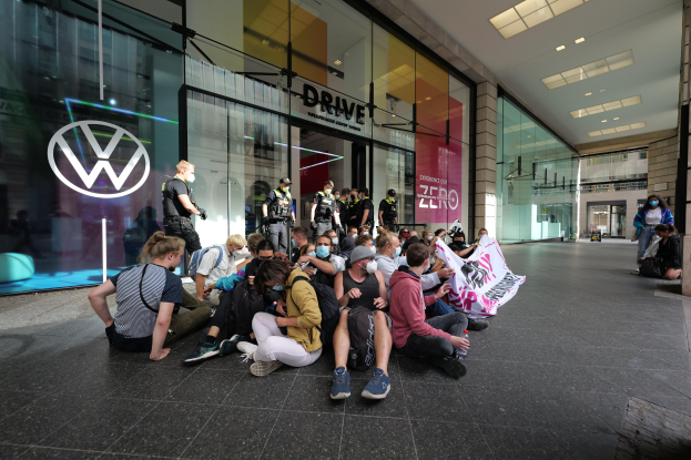 Eine Gruppe von Menschen sitzt vor einer Volkswagen-Verkaufsstelle mit Glaswänden auf dem Boden und hält ein Transparent mit Text.