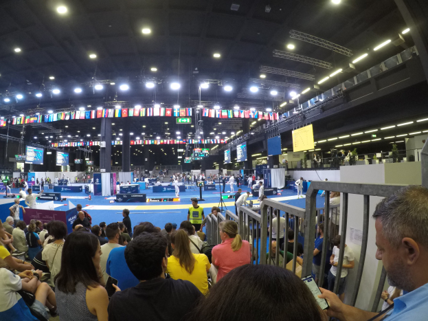Eine große Menschenmenge sitzt und steht in einer Indoor-Sportarena, mit Metallzaun auf der rechten Seite, Bildschirmen, Fahnen und Deckenleuchten im Hintergrund.