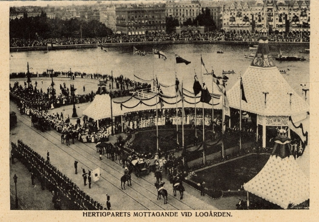 Ein altes Schwarz-Weiß-Foto einer Menge, die sich um einen Fasching mit Zelten, Fahnen, Stangen und Pferden im Vordergrund und Gebäuden, Bäumen und Wasser im Hintergrund versammelt hat, mit einem Text am unteren Bildrand.