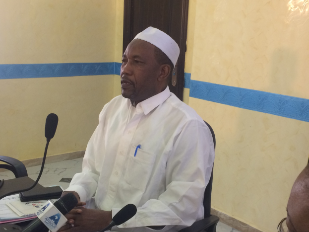 Ein Mann in einem weißen Kleid und Hut sitzt an einem Tisch mit Mikrofonen und spricht, während Papiere und Gegenstände auf dem Tisch sichtbar sind; ein weiterer Mensch mit Brille ist rechts von ihm, mit einer Wand und einer Tür im Hintergrund.