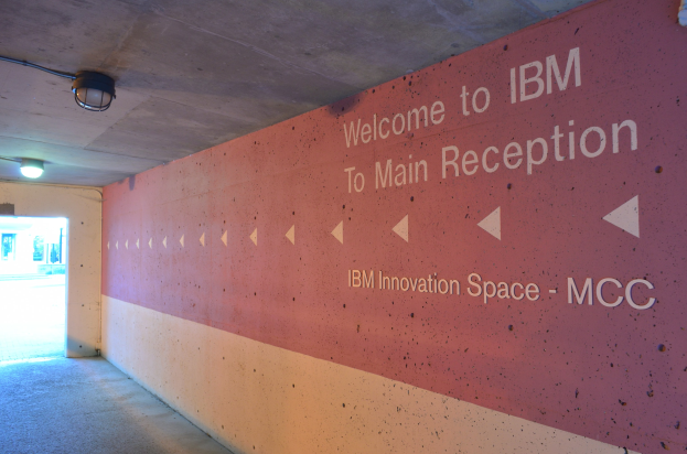 Eine Wand mit der Aufschrift "Willkommen bei IBM", beleuchtet von Deckenlampen und eine Tür im Hintergrund mit der Aufschrift "IBM Innovation Space MCC".