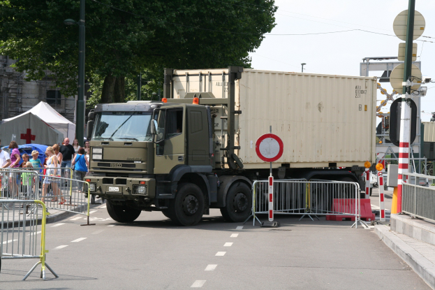 Ein Lkw parkt am Straßenrand, umgeben von Menschen, Absperrungen, Schildern, Straßenlaternen, Strommasten, Stromkabeln, Bäumen und Gebäuden unter einem klaren blauen Himmel.