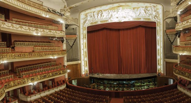 Großer Innenraum mit roter Gardine, Sitzplätzen, Geländern, Beleuchtung, Wandskulpturen und Bühnenobjekten, der an einen Konzertsaal erinnert.