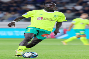 Ein Fußballspieler in gelbem T-Shirt und grünen Shorts ist im Einsatz auf dem Feld, mit einer blauen Tafel und unscharfen Figuren im Hintergrund.