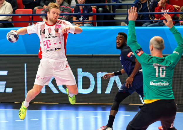 Ein Mann in einem pinken Hemd mit einem Fußball, mit der Hand eines anderen Menschen auf der linken Seite und ein paar Menschen im Hintergrund auf Stühlen sitzend.