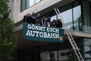 Eine Gruppe von Menschen auf dem Dach eines Gebäudes stehend, eine Fahne haltend, mit einem Baum links und Glasfenstern im Hintergrund, wahrscheinlich protestierend im Namen der deutschen Regierung.