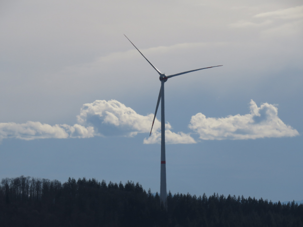Windkraftanlage im Wald mit bewölktem Himmel und Hügel im Hintergrund.