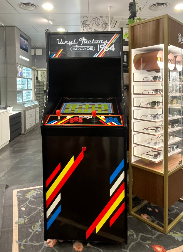 Ein Geschäft mit einem zentralen Retro-Arcade-Automaten, einer Brillen-Display-Ständer auf der rechten Seite und einer retro-stylischen Beleuchtung mit Schränken, Fernseher und anderen Gegenständen im Hintergrund.