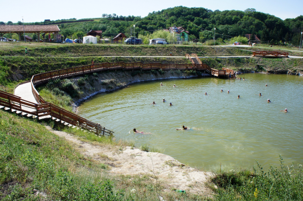 Gruppe von Menschen, die in einem Gewässer schwimmen, mit üppiger grüner Umgebung, einer Brücke mit Stufen, die ins Wasser führen, Hütten, Fahrzeugen, Pfählen und einem klaren blauen Himmel im Hintergrund.
