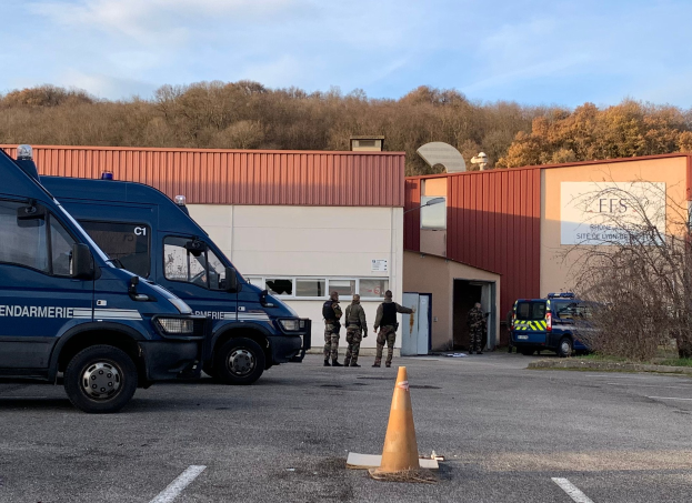 Polizeibeamte in Uniform stehen vor einem Gebäude mit Fenstern und Türen, mit Fahrzeugen und einem Verkehrskegel auf der Straße, Bäumen im Hintergrund und einem bewölkten Himmel.