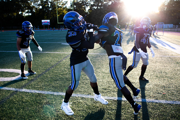 Gruppe junger Männer beim Fußballspielen auf einem Feld mit Bäumen und einem klaren blauen Himmel, die Helme und Stollenschuhe tragen.