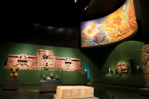 Ein Museumsausstellungsraum mit verschiedenen Artefakten, darunter Skulpturen, Gegenstände auf Tischen, eine Texttafel, eine Wandmalerei und Deckenbeleuchtung im Anthropologie-Museum von Mexiko-Stadt.