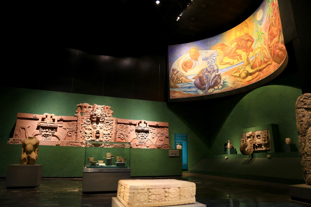 Ein Museumsausstellungsraum mit verschiedenen Artefakten, darunter Skulpturen, Gegenstände auf Tischen, eine Texttafel, eine Wandmalerei und Deckenbeleuchtung im Anthropologie-Museum von Mexiko-Stadt.