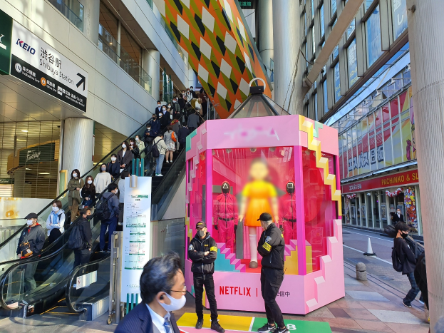 Eine Gruppe von Menschen steht vor einem modernen Gebäude mit Glaswänden und Texttafeln, einige tragen Masken, mit einer Rolltreppe im Blick; ein pinker Kasten in der Mitte zeigt Schaufensterpuppen und trägt 'Netflix' neue Netflix-Bühne in Tokio'.