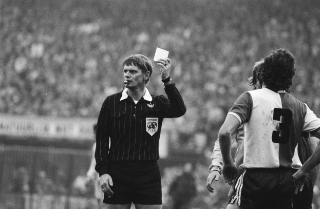 Schwarzes und weißes Bild eines Schiedsrichters mit einer roten Karte vor einer Menge von Füßballspielern, mit einem Zaun im Hintergrund.