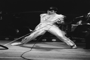 Elvis Presley performs on stage at the Apollo Theater in Las Vegas, wearing a white jumpsuit and holding a microphone, with another musician to his right and stage lights in the background.