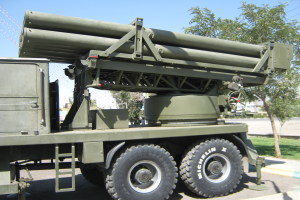 Ein militärisches Lastwagenfahrzeug mit einer Rakete fährt auf einer Straße, umgeben von Gras, Bäumen, Gebäuden und einem Maschendrahtzaun.