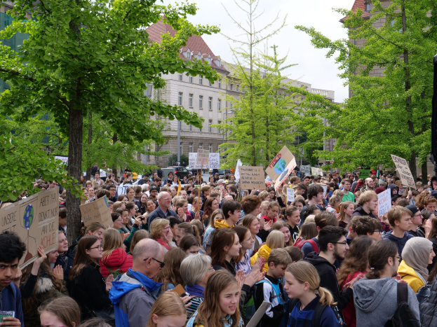 Eine große Menge von Menschen protestiert vor einem Gebäude in Berlin, mit Schildern, Bäumen, Fahrzeugen, einem Lautsprecher und dem Himmel.
