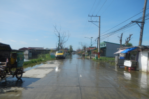 Überflutete Straße mit einem Rikscha auf der Seite geparkt, Fahrzeuge fahren, Strommasten mit Drähten, Gebäude und Bäume im Hintergrund, sichtbarer Himmel und ein Wasserzeichen in der rechten unteren Ecke.