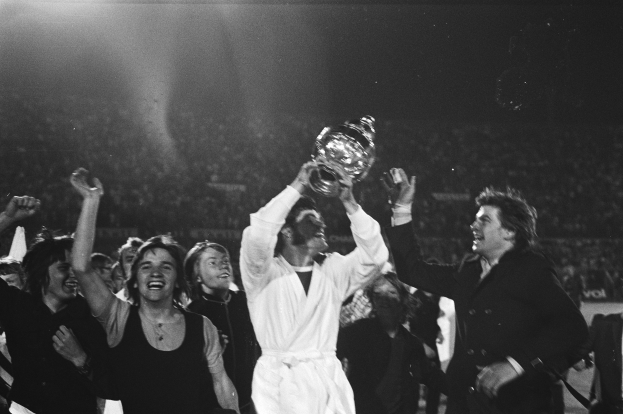 Schwarzes und weißes Bild einer Gruppe von Menschen, die mit einem Pokal feiern, wobei die zentrale Figur ihn in einem hell erleuchteten Stadion hochhält.