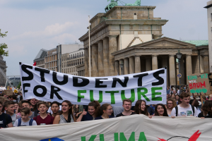 Gruppe von Schülern marschiert in Berlin mit einem bunten "Students for Future"-Schild gegen eine Kulisse aus Gebäuden, Bäumen und Himmel.