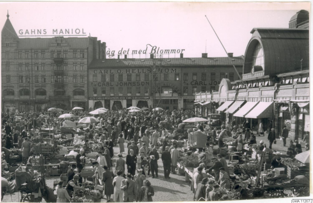 Schwarz-weiß-Foto eines belebten Berliner Markts mit Menschen, Gemüsewagen und Gebäuden mit Fenstern im Hintergrund.