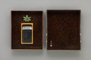 Eine schwarze und goldene rechteckige Zigarettenbox mit glänzender Oberfläche, die ein leuchtend grünes Marihuana-Blatt in der Mitte zeigt, auf einer grauen Oberfläche.