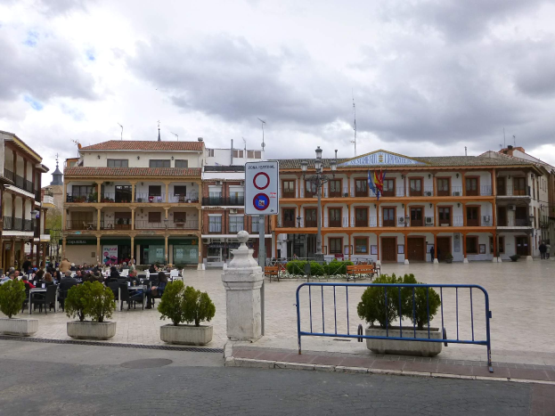 Ein belebter Stadtplatz mit Menschen auf Stühlen sitzend und stehend, umgeben von Topfpflanzen, Metallabsperrungen, Straßenlaternen mit Flaggen, einem Schild, Gebäuden mit Fenstern und einem bewölkten Himmel.