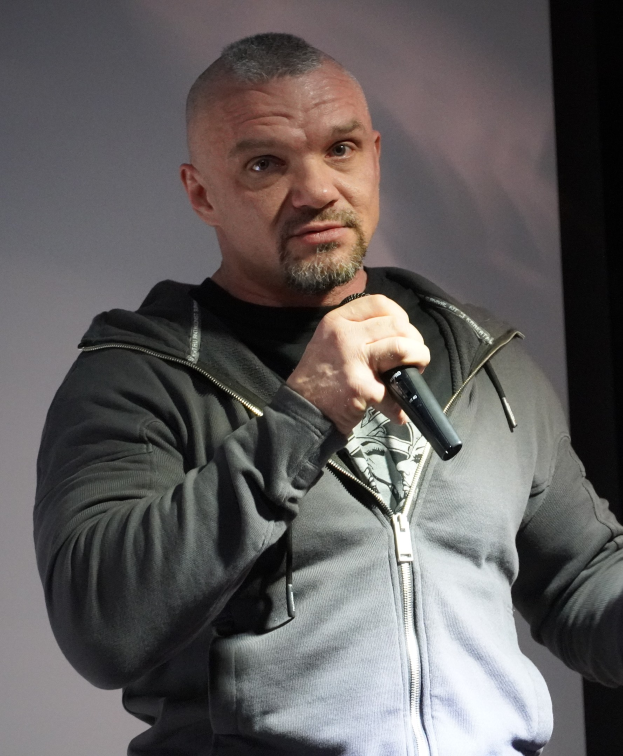 Komiker in schwarzem Anzug und weißem Hemd, der ein Mikrofon hält und selbstbewusst vor einer Wand steht.