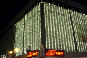 Ein Gebäude in Berlin, Deutschland, geschmückt mit festlichen Weihnachtslichtern, umgeben von Bäumen und Laternenmasten, mit Texttafeln in der Nähe und dem Himmel im Hintergrund.
