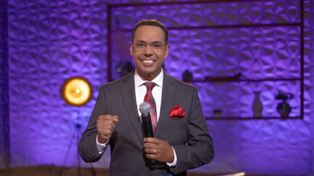 Stephen Colbert, Moderator von *The Daily Show with Trevor Noah*, steht auf einer Bühne und hält ein Mikrofon, trägt einen Anzug, Krawatte und Brille, mit einer Lampe und Ständer im Hintergrund.