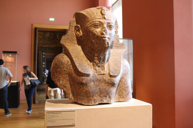 Ein Büste eines ägyptischen Pharaos wird in einem Museum mit einer Texttafel, einer Gruppe von Menschen, einer Frau mit einer Tasche, einer Tür, einer Wand und einer Decke mit Lampen gezeigt.