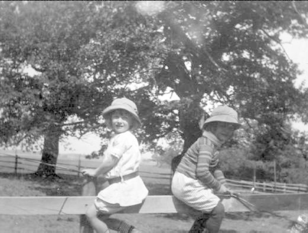 Zwei junge Jungs in Mützen stehen auf Gras und halten Baseballschläger, mit Bäumen und einem Zaun im Hintergrund, auf einem Schwarz-Weiß-Foto.