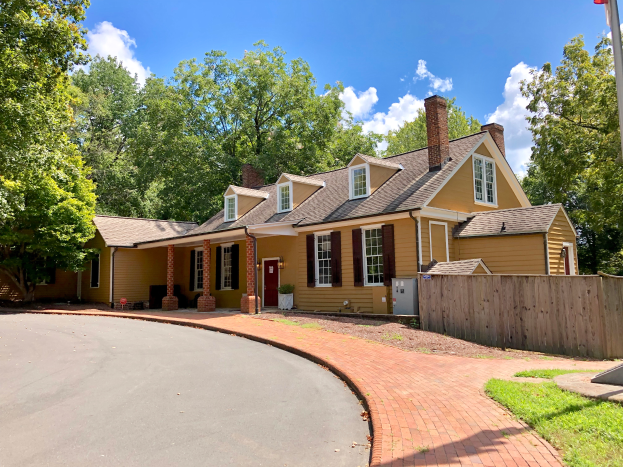 Ein gelbes Haus mit einem Backsteinweg, einem hölzernen Zaun und grünem Gras, das sich an 604 W Main Street in Charlotte, NC 28277 befindet, mit einem Pfahl im Vordergrund, Bäumen im Hintergrund und einem klaren blauen Himmel darüber.