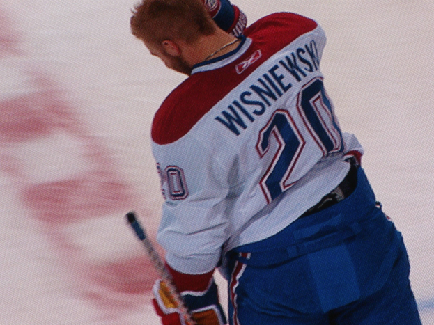 Ein Eishockeyspieler in einem "Wisniewski"-Trikot steht auf dem Eis und hölt einen Schläger, mit einem konzentrierten und entschlossenen Gesichtsausdruck.