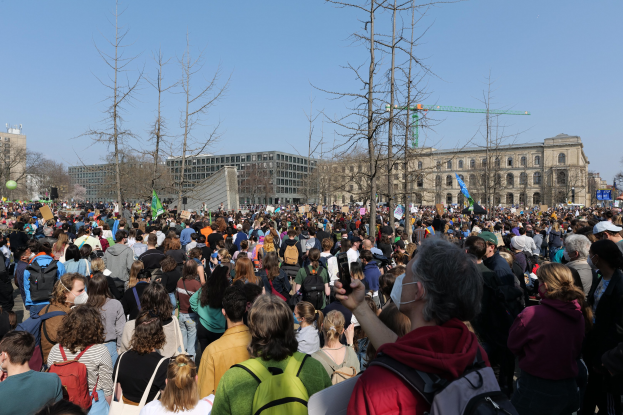 Große Menge von Menschen mit Taschen und Schildern vor einem Gebäude mit Bäumen und einem klaren Himmel, die an einer Klimaprotest teilnehmen.