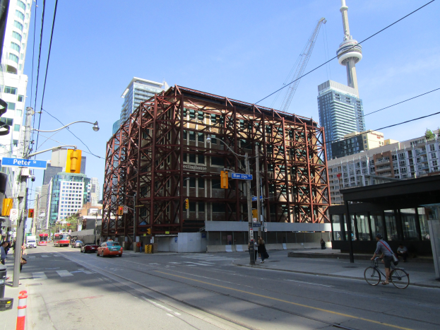 Eine belebte Stadtstraße mit einem großen Baugerüst im Hintergrund, Fahrzeuge auf der Straße, eine Person auf einem Fahrrad und Fußgänger auf dem Gehweg, Ampeln, Pfosten mit Schildern, ein Kran und der Himmel.
