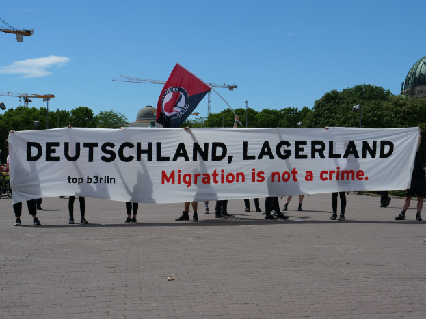 Eine Gruppe von Menschen mit einem Transparent, auf dem "Deutschland, Lagerland Migration ist kein Verbrechen" steht, und einer Flagge, im Hintergrund Straßenmasten, Laternen, Bäume, Kräne, ein Gebäude und ein bewölkter Himmel.