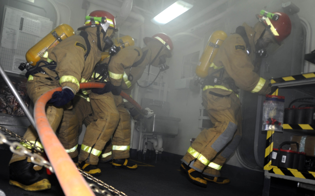 Feuerwehrleute in Schutzausrüstung arbeiten an einem Feuerlöschfahrzeug mit Ausrüstungsgestellen und Hintergrundinfrastruktur.