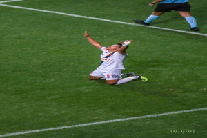 Ein Fußballspieler kniet auf dem Boden und feiert mit ausgestreckten Armen, trägt eine Sportuniform und Schuhe.