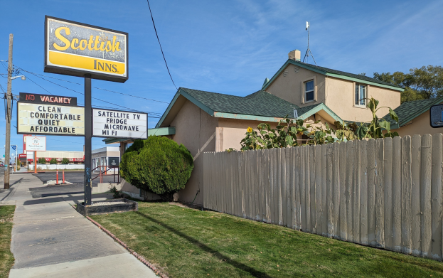 Ein schottisches Motel in San Francisco, Kalifornien, mit einem Haus mit Dach und Fenstern, einem Holz-Zaun, Pflanzen, Gras, Bäumen, Schildern, Strommasten mit Drähten, einer Straßenlaterne, Fahrzeugen auf der Straße und einer bewölkten Himmel.