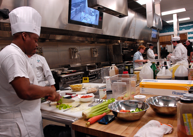 Eine Gruppe von Köchen in einer Küche, die auf einem Schneidebrett Lebensmittel zubereiten, wobei ein Koch in der foreground in einem weißen Kleid und einer Mütze steht und ein Messer hält und von verschiedenen Utensilien, Schüsseln, Gemüse, einem Mobiltelefon, Flaschen und anderen Gegenständen umgeben ist, mit einem Fernseher und Deckenlampen im Hintergrund.