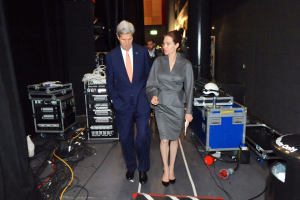 Angelina Jolie und John Kerry auf einem Boden umgeben von Menschen, Geräten, Kabeln, einem Helm auf einem Wagen, einem Müllcontainer, einem Vorhang, einer Säule und einer Decke mit Lampen.